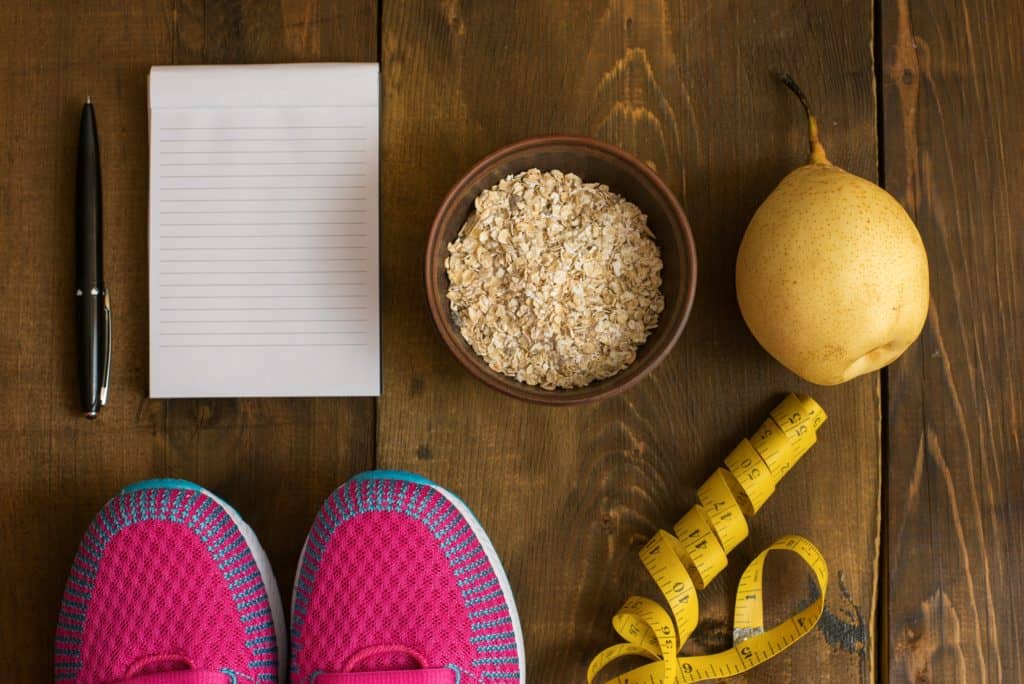 Running shoes, measuring tap, notepad, oatmeal, and a pear implying a narrative of weight loss